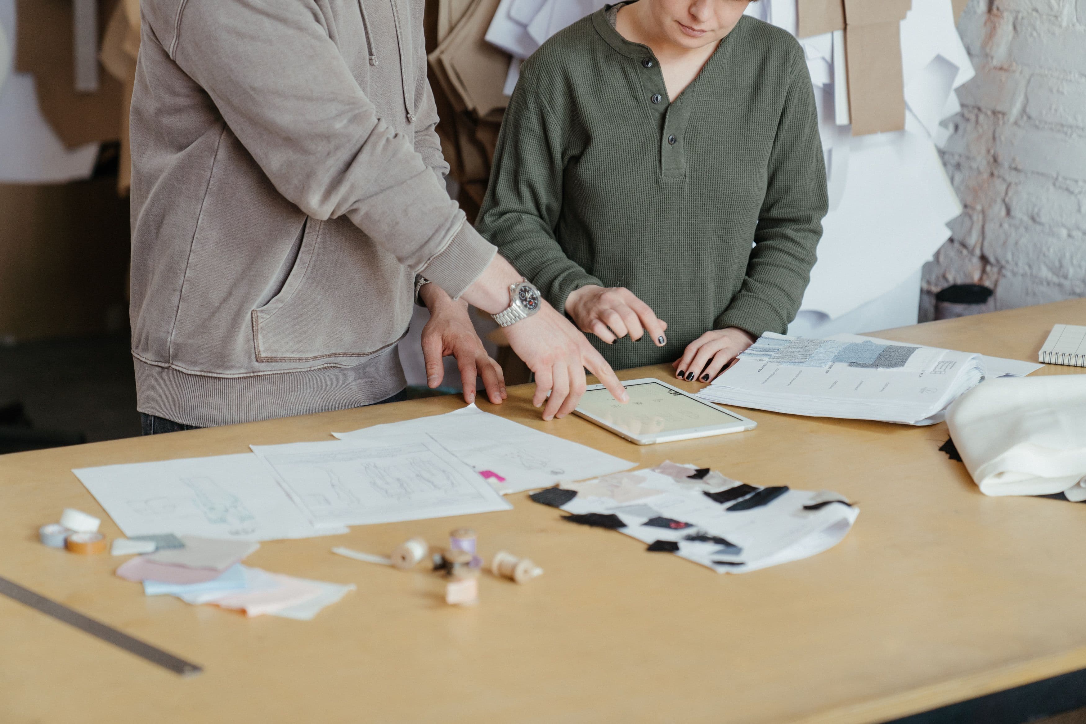 Hands reviewing garment designs on a tablet beside fabric samples and patterns
